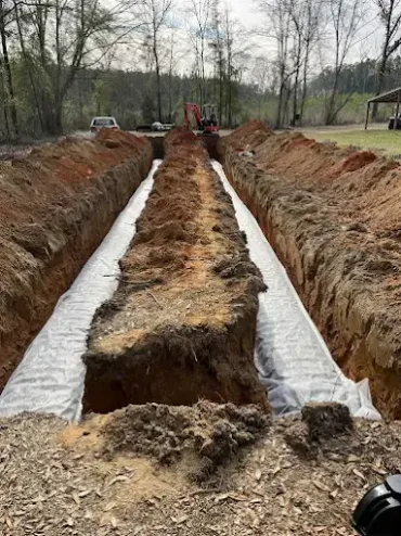A wide, outdoor trench construction site featuring two parallel, fabric-lined drainage channels with a dirt wall between.