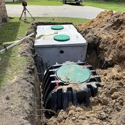 A concrete septic tank and a black plastic pump chamber installed in a narrow, freshly excavated dirt trench.