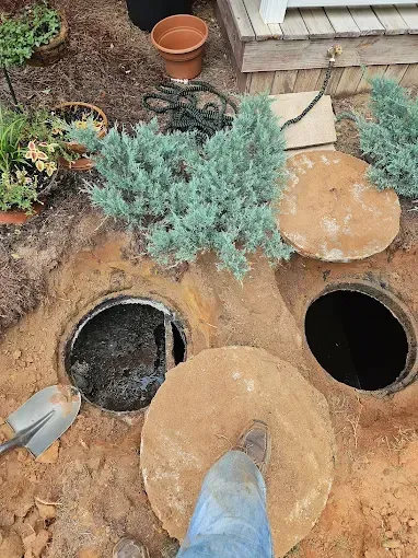 A person stands on a concrete lid next to an open septic tank riser in a dirt-filled yard with decorative shrubs.