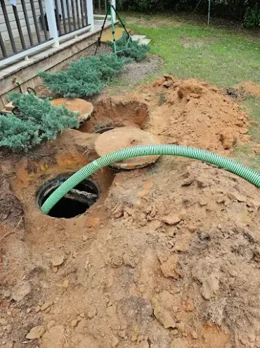 A green hose is inserted into an open, excavated septic tank access hole near a house foundation and landscaping.