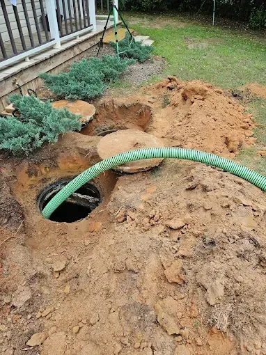A green suction hose inserted into an open, excavated septic tank access port near a house foundation.