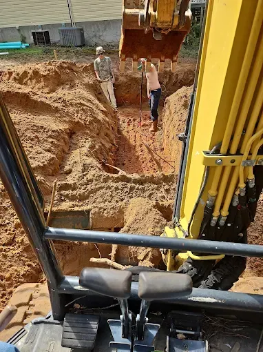 View from inside an excavator cab looking down at two people standing in a deep, dirt-filled construction trench.