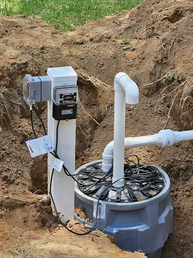 An installed outdoor sump pump basin with electrical control box and white PVC plumbing in an excavated dirt trench.