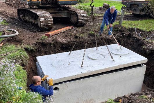 Two workers in a trench guide a concrete septic tank lid lowered by an excavator into a hole in the ground.