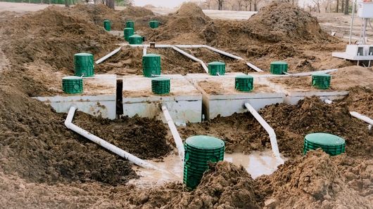 A cluster of underground septic tanks with green riser lids and white PVC pipes, shown during installation in a dirt trench.