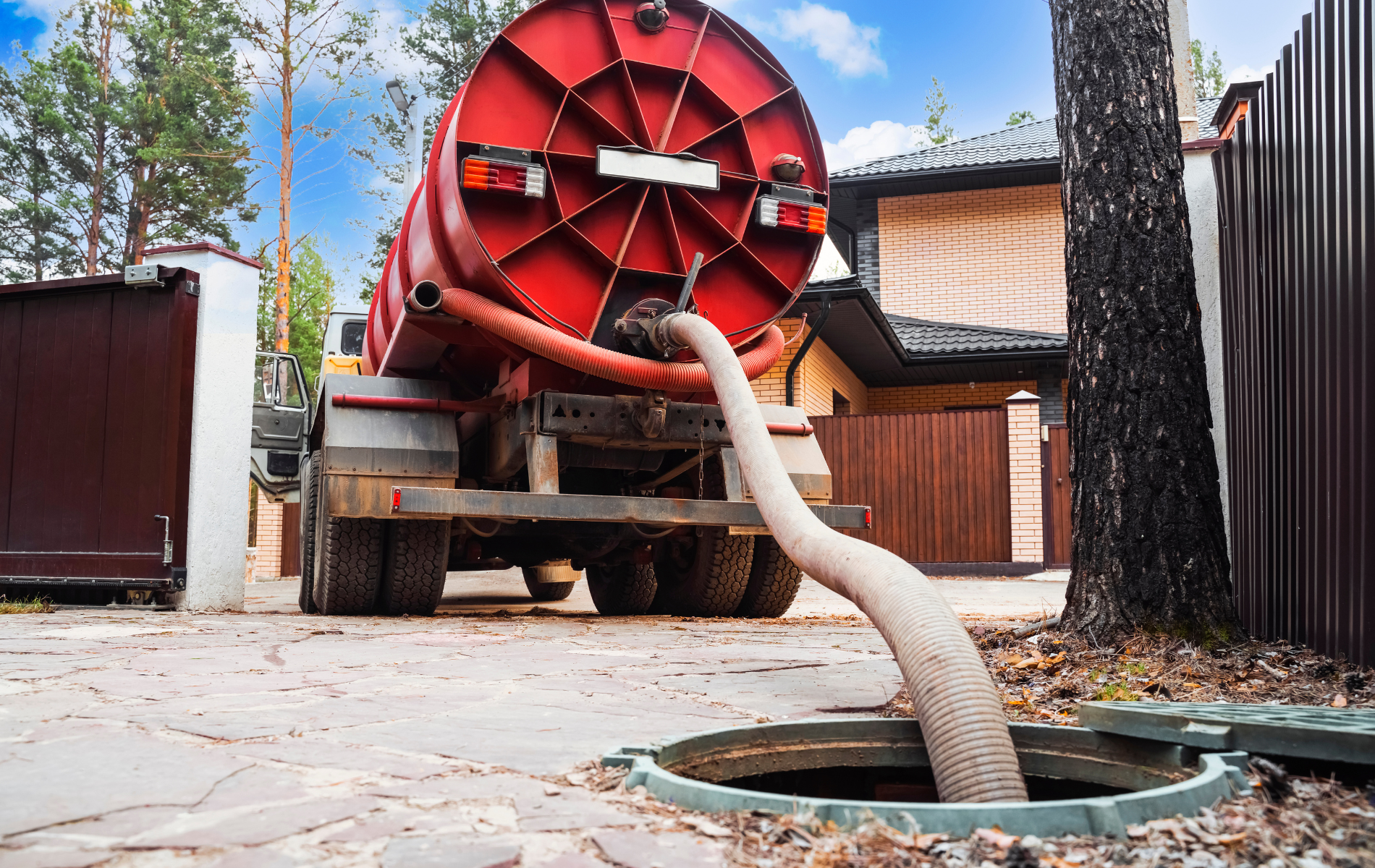 A red vacuum truck parked in a residential driveway is pumping sewage from an open ground-level septic tank access port.