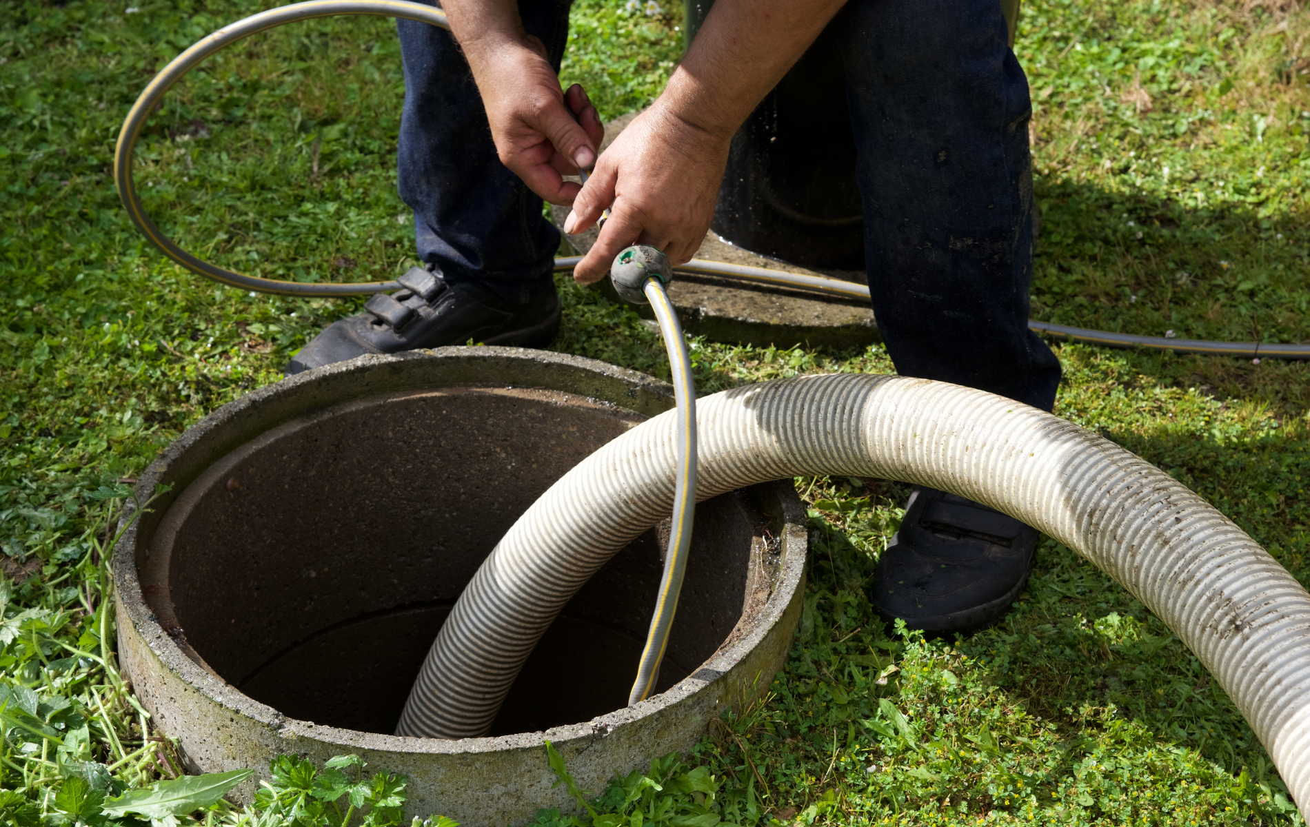 A person uses a vacuum hose to pump out a septic tank in a grassy yard.