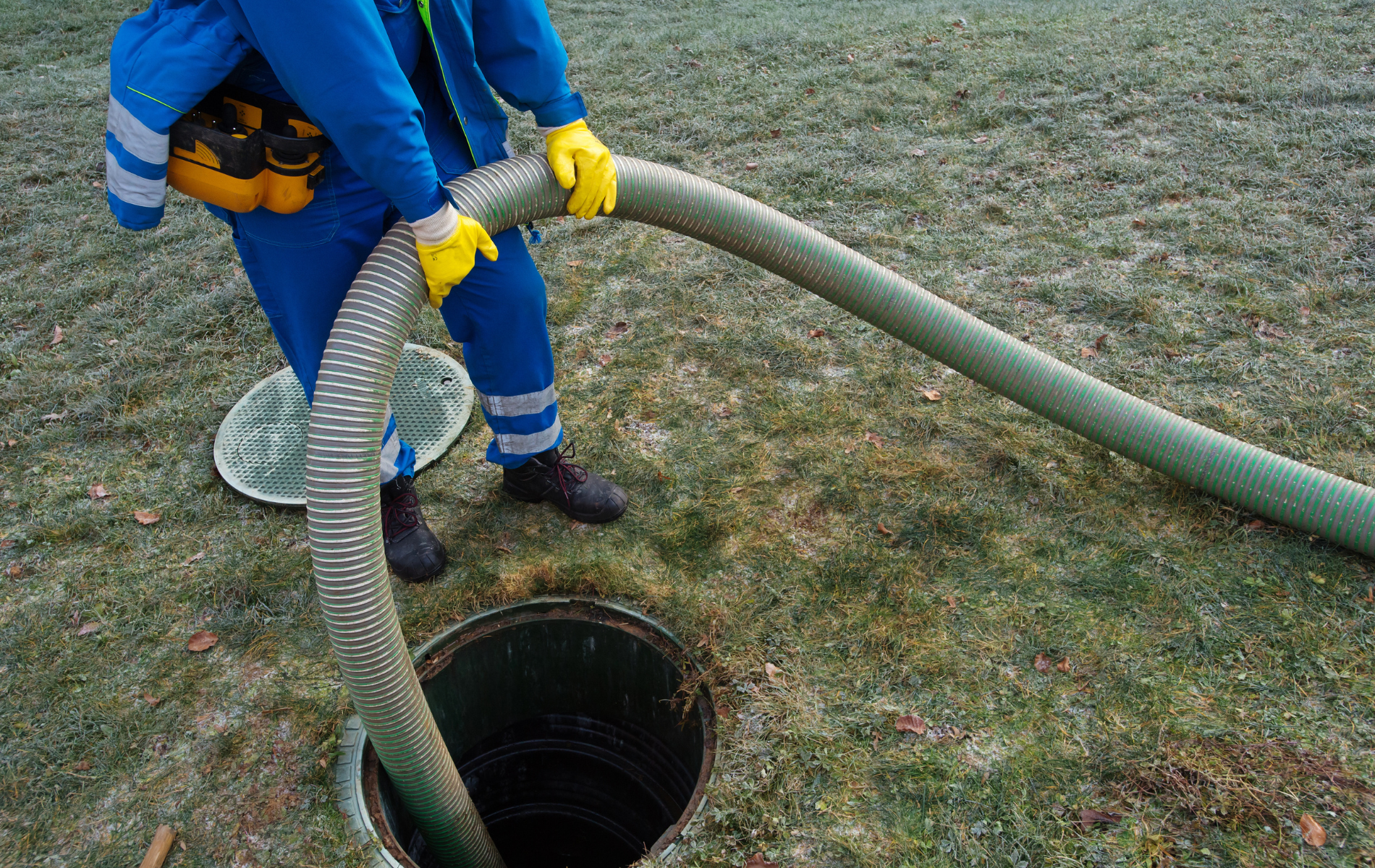A worker in blue coveralls and yellow gloves uses a large, flexible hose to clean out an open utility access manhole.
