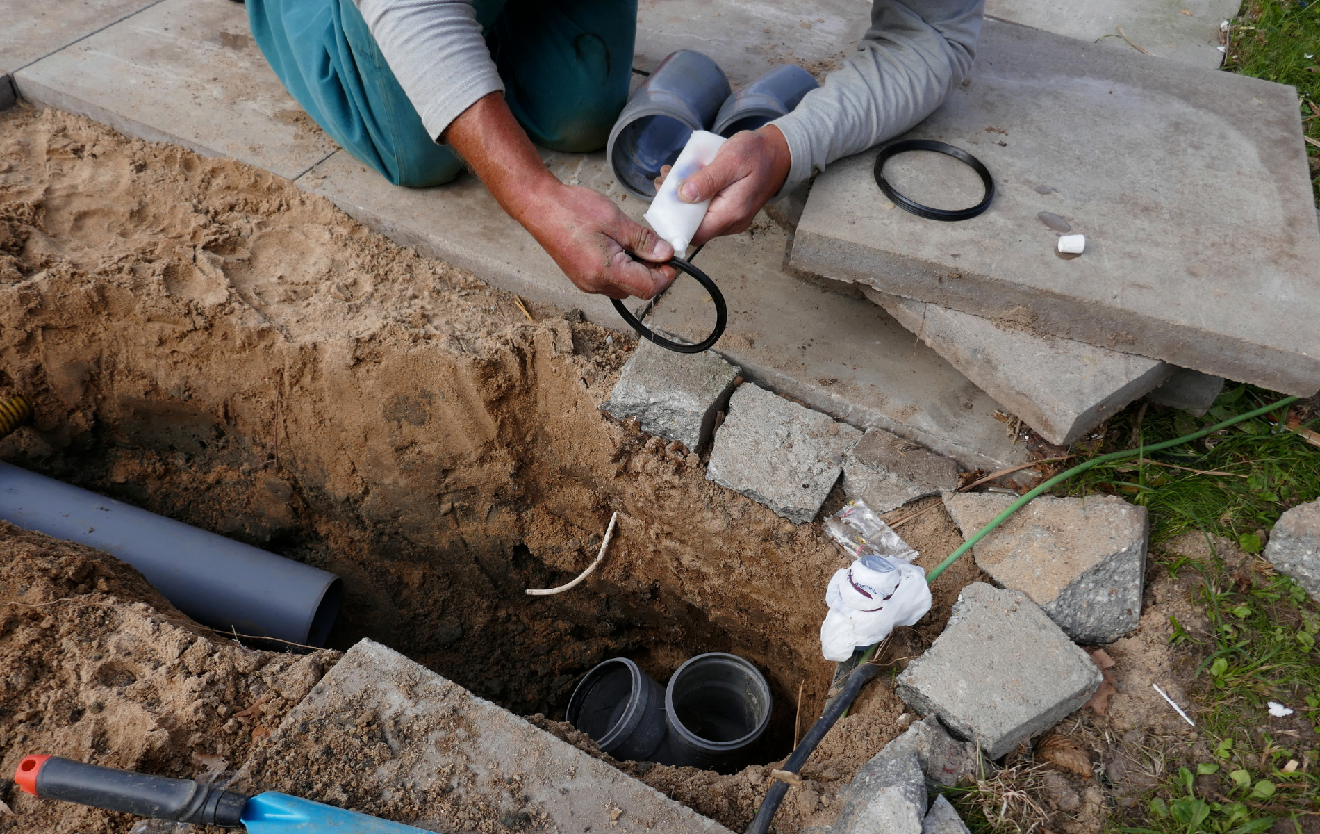 A person kneels by a narrow dirt trench, fitting a black rubber O-ring seal onto a gray plastic plumbing pipe fitting.