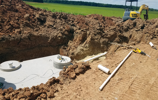 A concrete septic tank installed in a deep dirt excavation pit with white PVC pipes laid nearby and a small excavator.