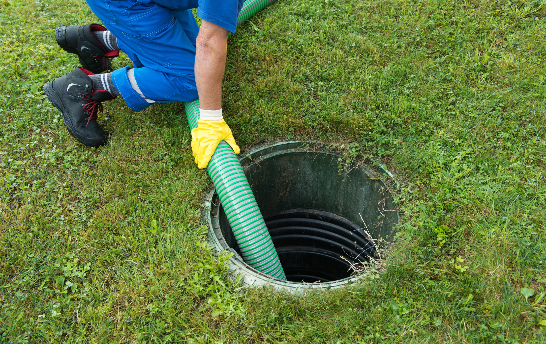 A person in blue workwear and yellow gloves inserting a green vacuum hose into a black septic tank in a grassy yard.