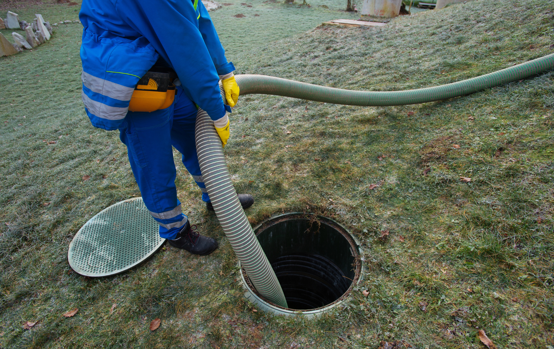 A worker in a blue uniform uses a large hose to pump out a septic tank in a grassy yard.