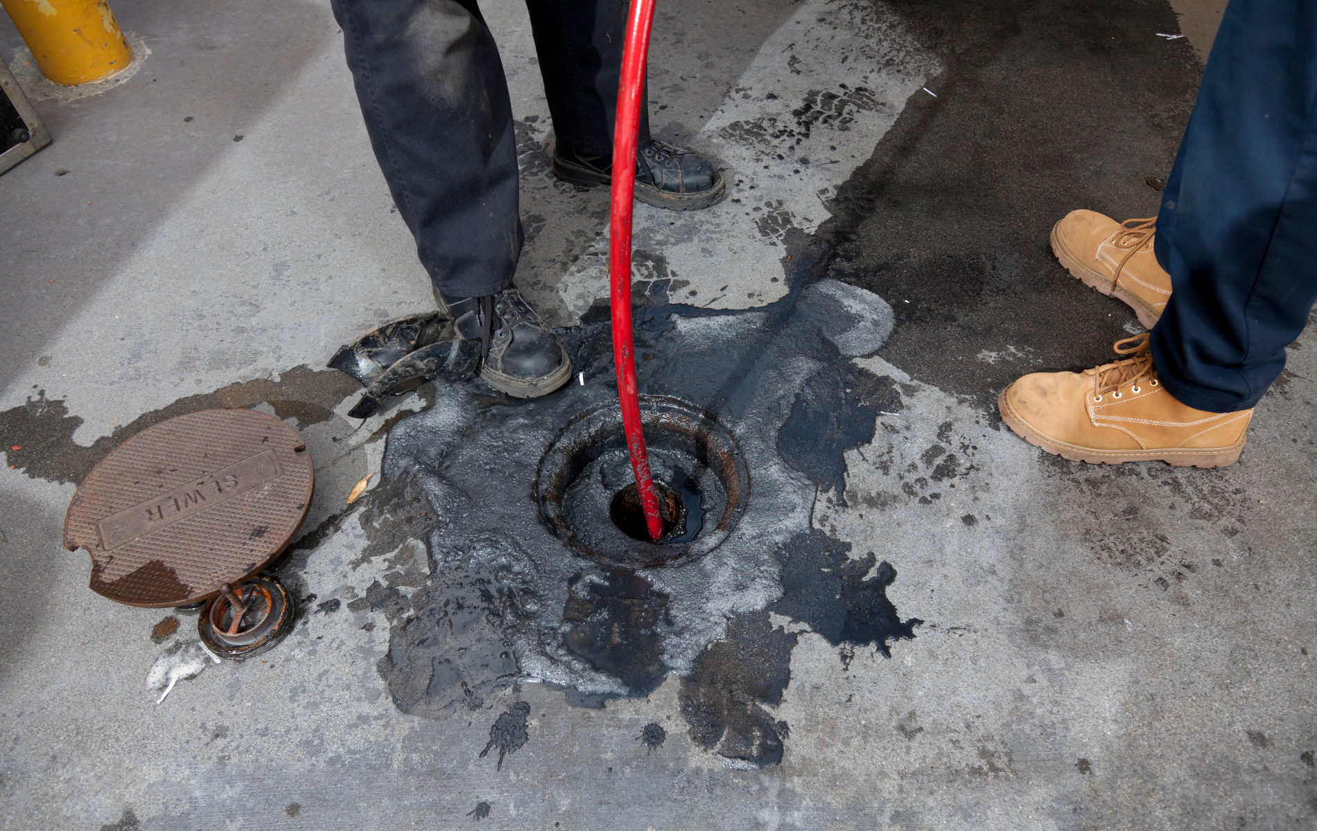 Two people stand by a manhole cover on a concrete surface, with a red hose inserted into the open drain.