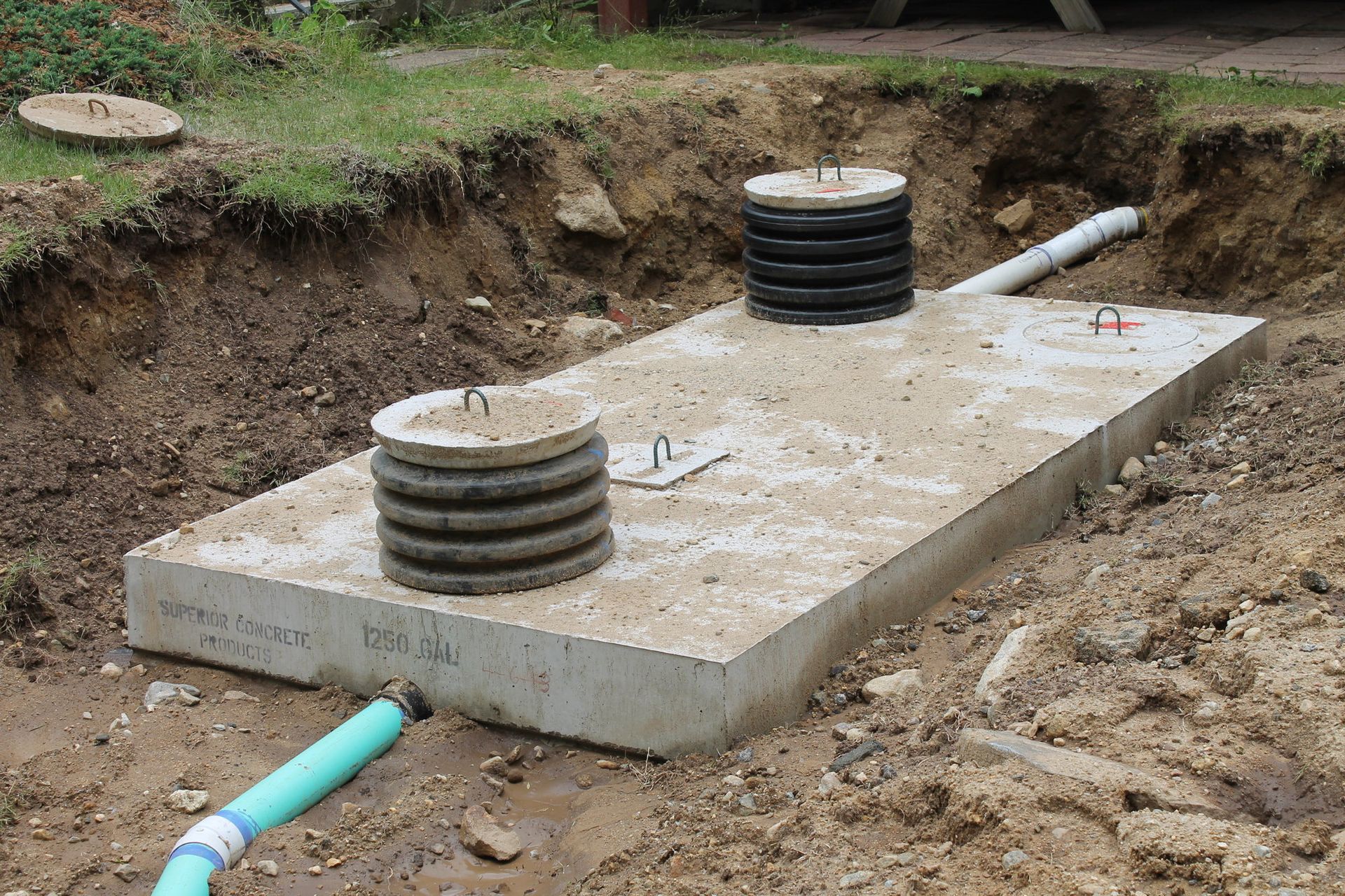 A rectangular concrete septic tank installed in a dirt excavation, featuring two access risers and connected drainage pipes.