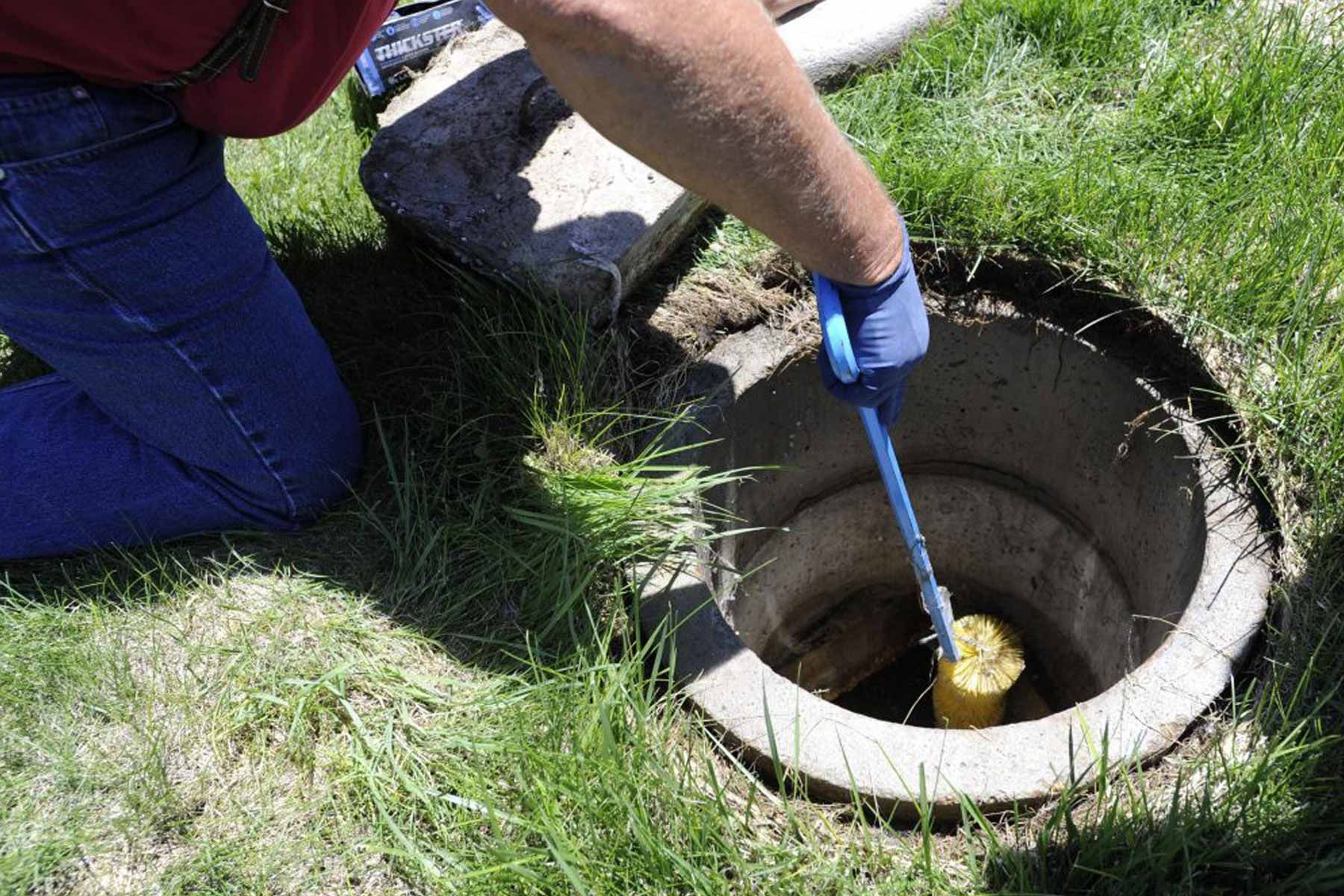 A person kneeling on grass uses a long-handled tool to retrieve a yellow cylindrical object from an open concrete pipe.