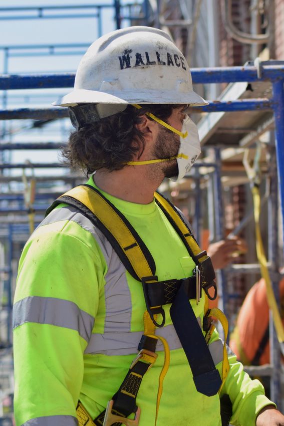 Construction worker in a safety harness and hard hat, wearing a mask. Yellow safety vest and scaffolding in background.