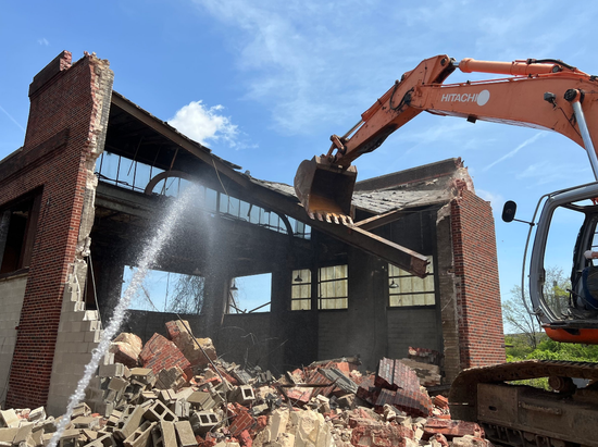 Excavator demolishing a brick building; water sprays to control dust under blue sky.