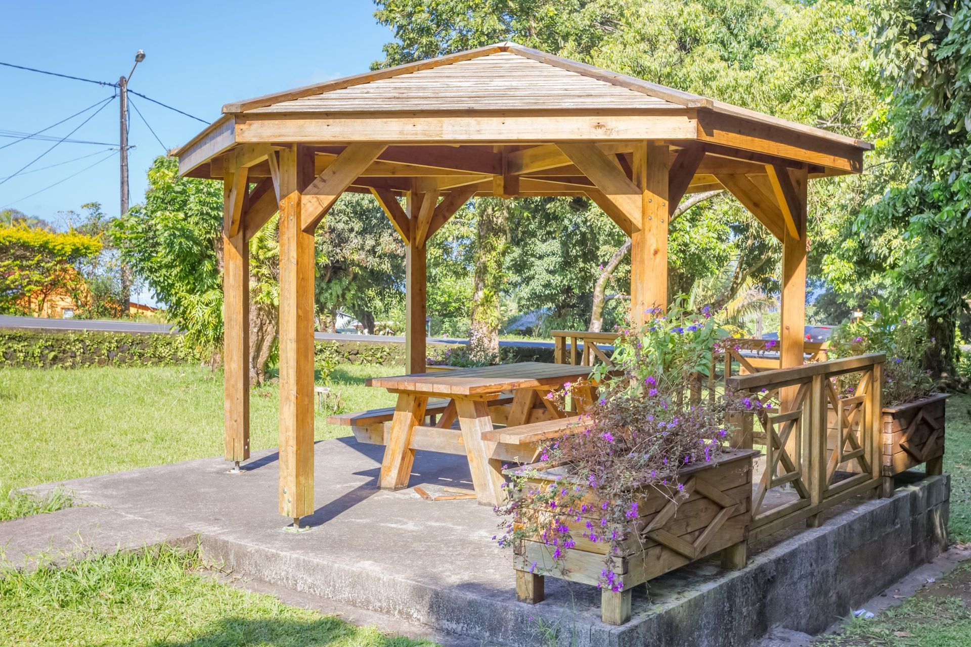 Wooden gazebo with picnic table, surrounded by greenery, on a concrete base
