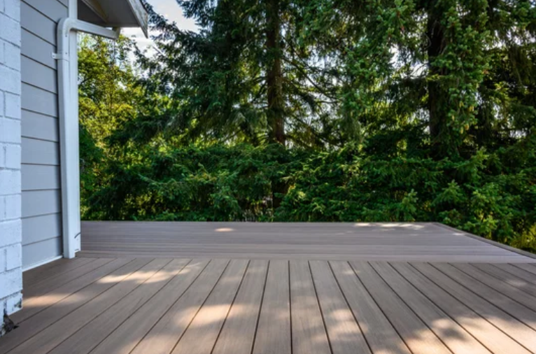 A brown composite deck next to a gray house with lush green trees in the background