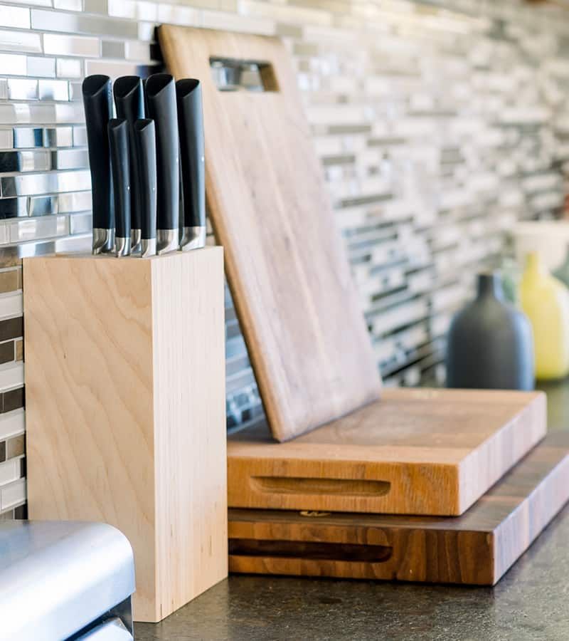 Knife block with knives, wooden cutting boards on a countertop, against a backsplash