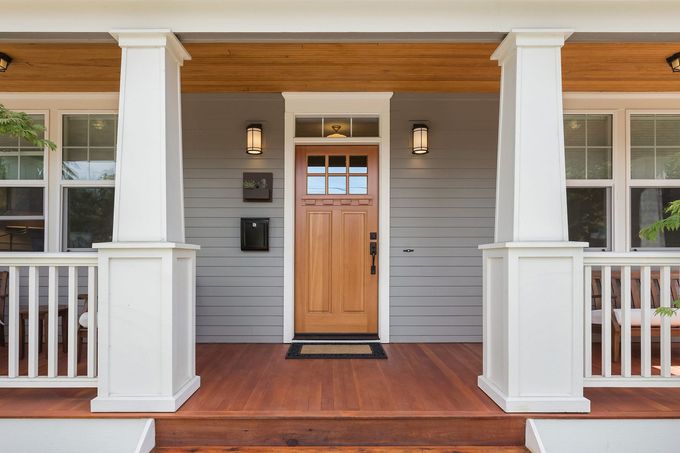 A wooden front door under a porch with white columns, gray siding, and a wood floor.