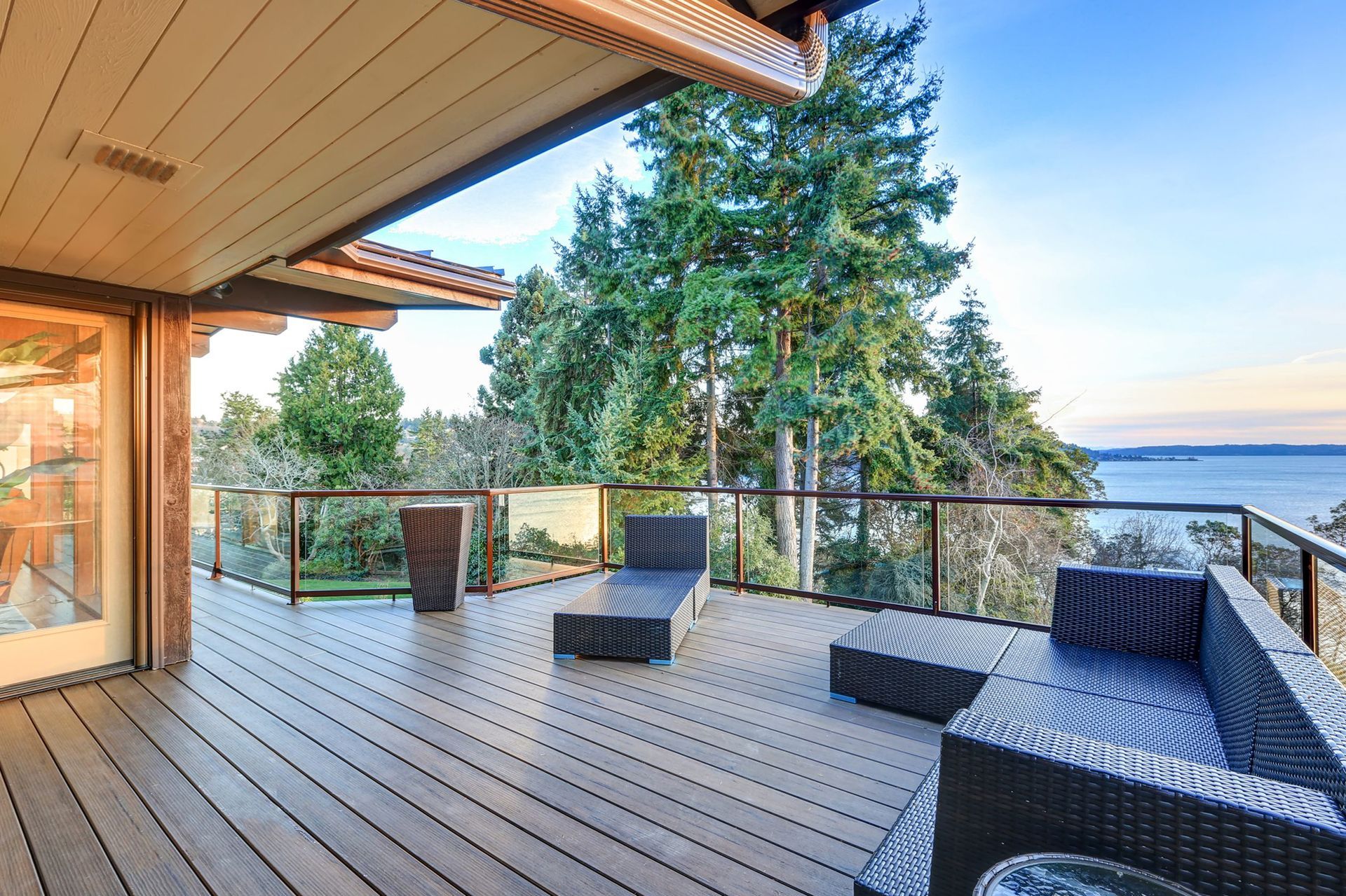 Wooden deck overlooking water, furnished with black wicker furniture and glass railings, tall trees in the background