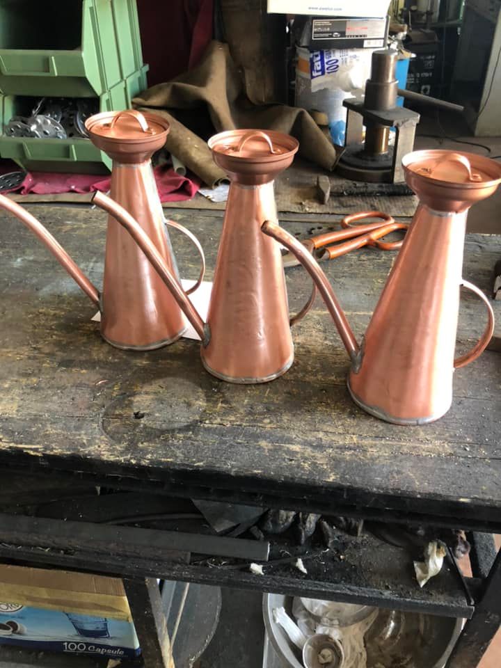 Su un banco da lavoro di legno scuro e consumato, tre annaffiatoi lucidi color rame con lunghi beccucci sono allineati in fila.