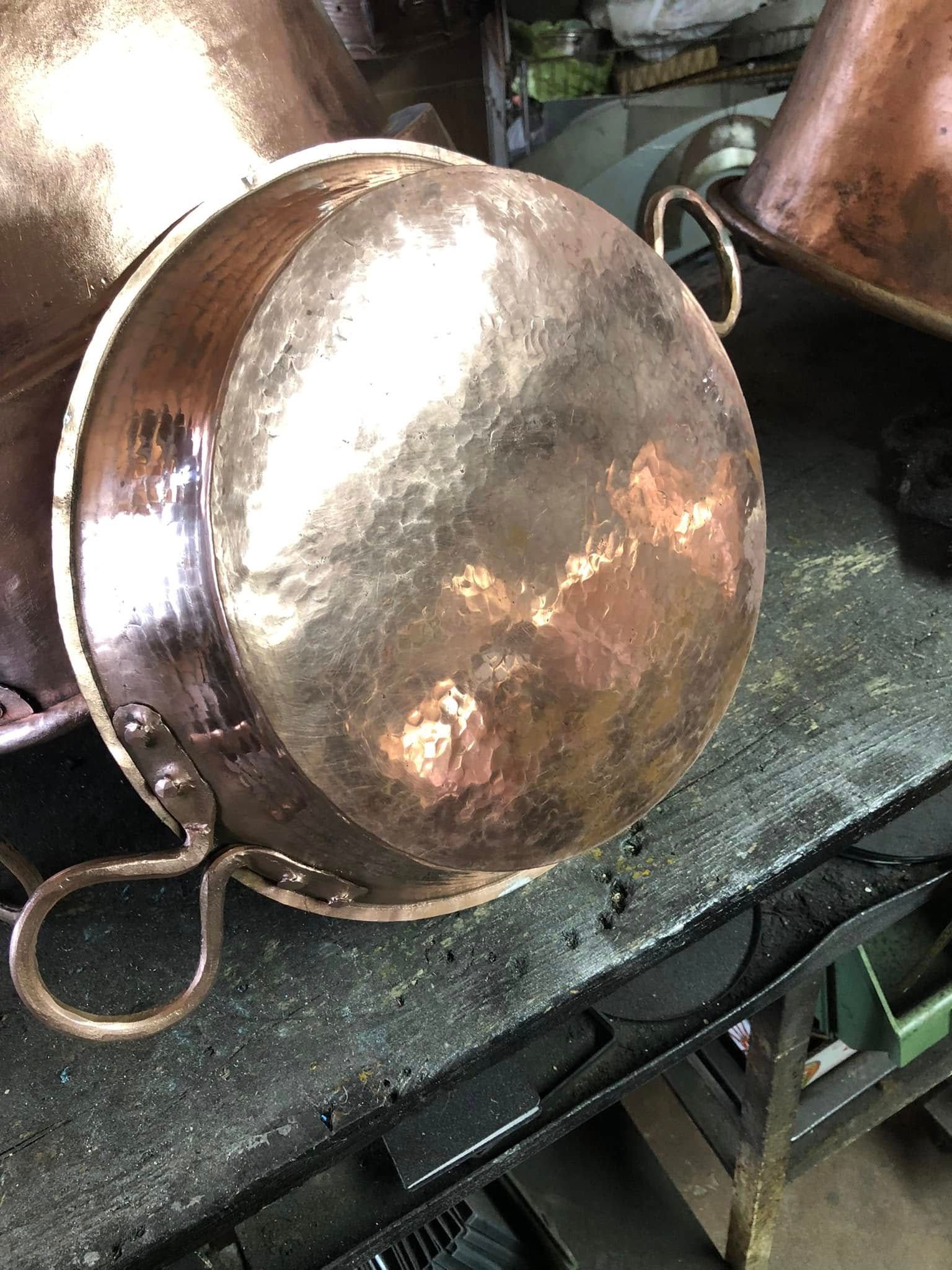 In un'officina, una pentola di rame con manici ad anello in metallo è appoggiata su un banco da lavoro scuro e ruvido.