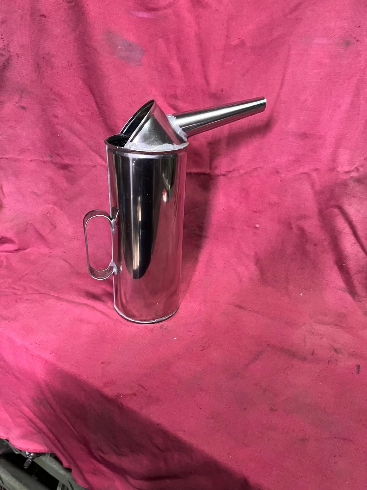 Una lattina d'olio in metallo argentato con beccuccio e manico, su uno sfondo di tessuto rosa.