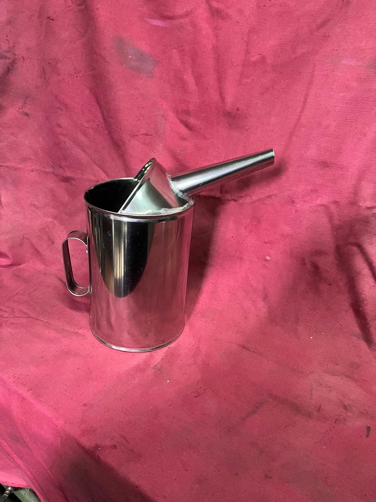 Una brocca per versare l'olio in acciaio inox color argento, con manico e beccuccio lungo e affusolato, su uno sfondo di tessuto rosa scuro.