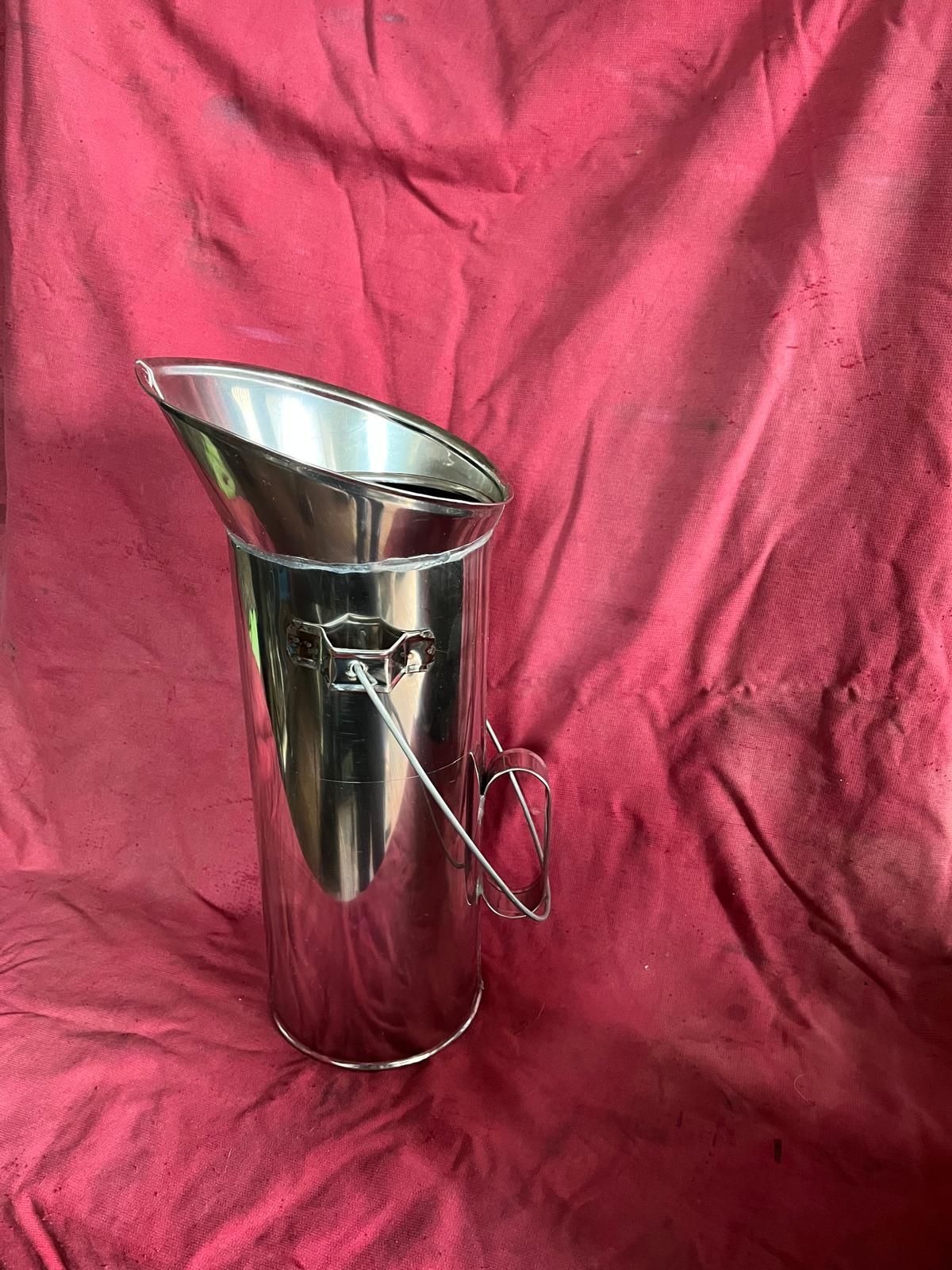 Una brocca alta e cilindrica in acciaio inox con apertura superiore inclinata, manico ad anello e manico laterale, rivestita in tessuto rosso.