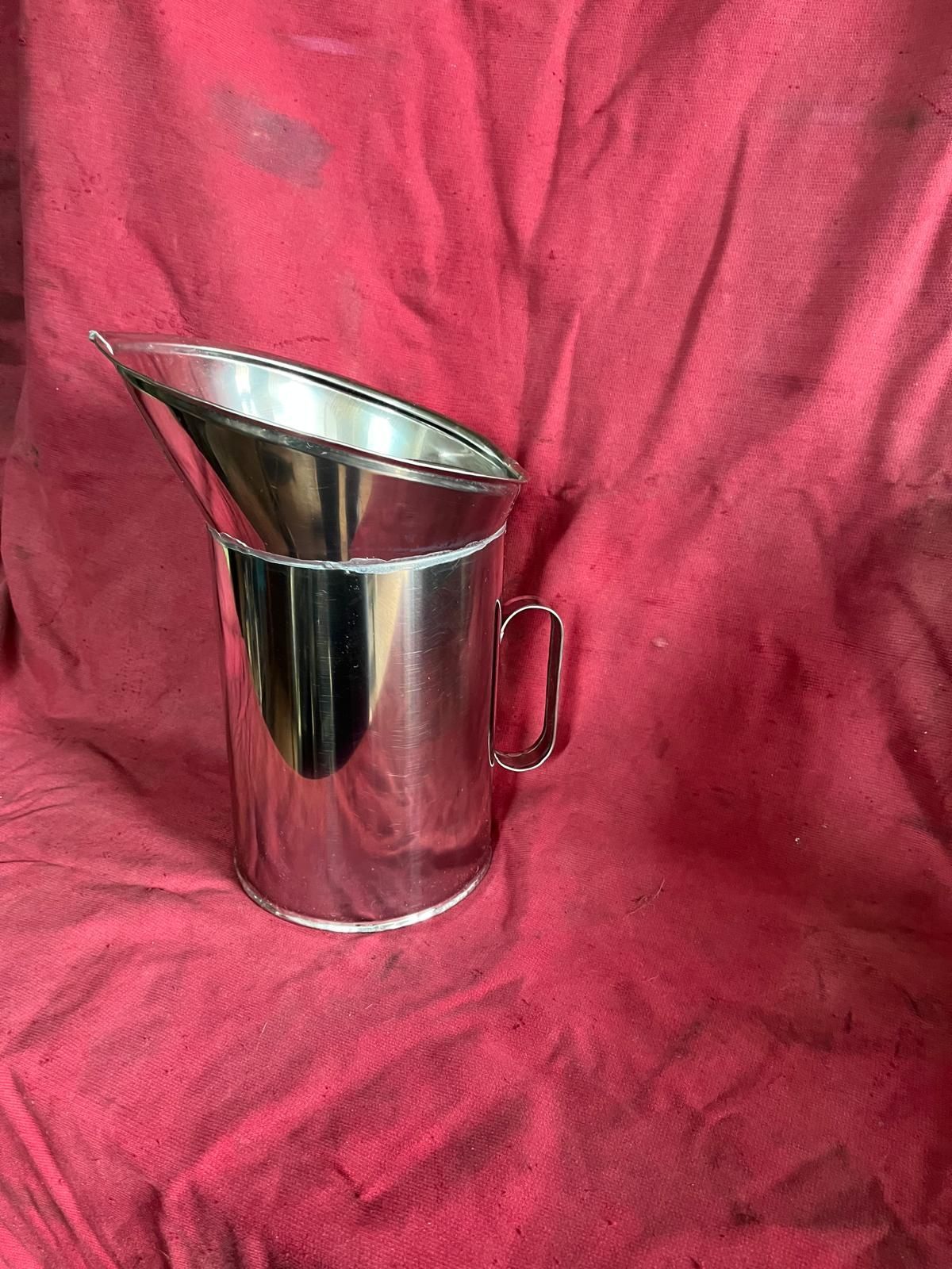 Una brocca in acciaio inossidabile con parte superiore inclinata e manico, centrata su uno sfondo di tessuto rosso.