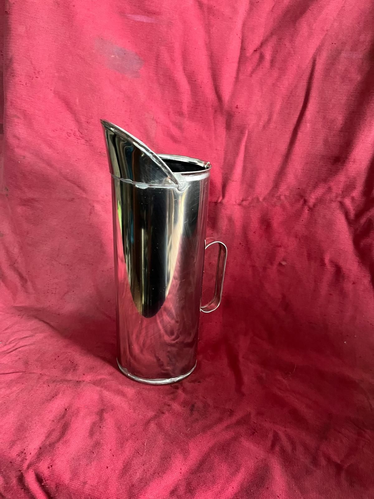 Una brocca cilindrica di metallo lucido con beccuccio e manico, appoggiata su uno sfondo di tessuto rosso scuro dalla trama marcata.