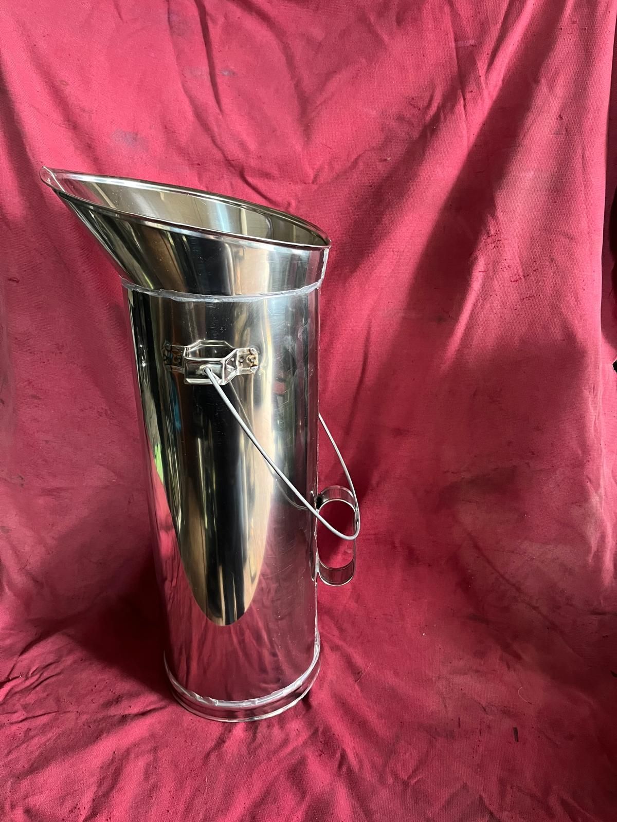 Una brocca cilindrica in acciaio inox lucido con beccuccio svasato e un piccolo manico in filo metallico su sfondo rosso.