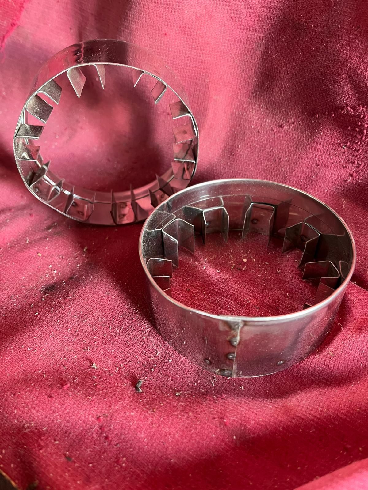 Due stampini per biscotti circolari in metallo con bordo interno smerlato e seghettato, poggiati su una superficie ruvida di colore rosa intenso.