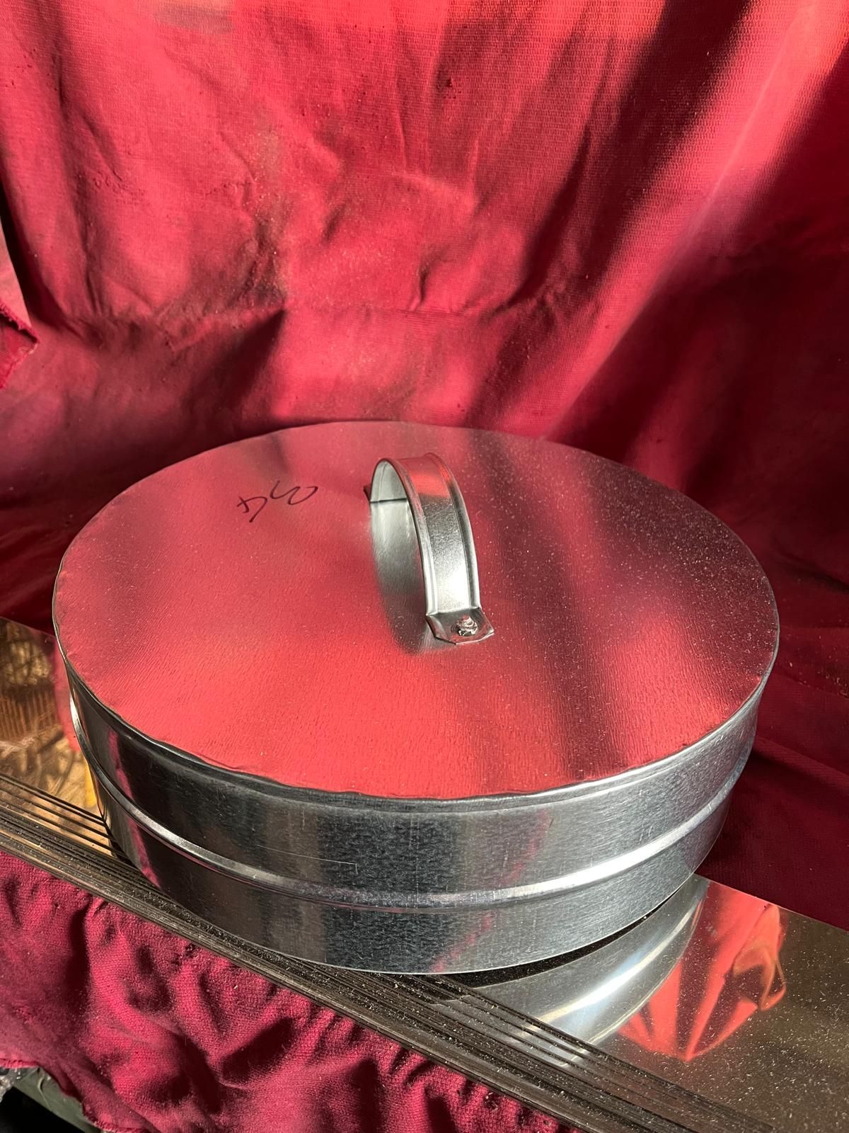 Un contenitore rotondo in acciaio zincato con coperchio piatto e maniglia centrale in metallo, appoggiato su una superficie di tessuto rosso.