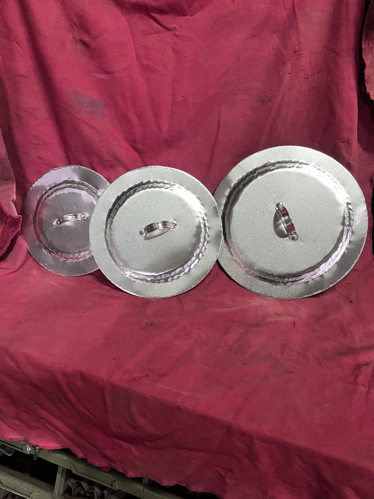 Tre coperchi metallici circolari di diverse dimensioni, disposti orizzontalmente su uno sfondo di tessuto drappeggiato color bordeaux.