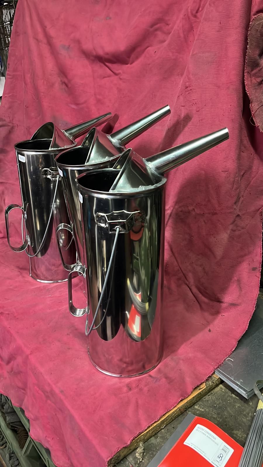 Tre taniche per versare l'olio in metallo lucido, complete di beccuccio e manico, disposte su una superficie di stoffa rossa.