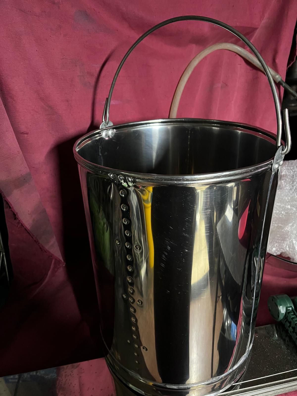 Un secchio in acciaio inox lucido con una cucitura verticale di rivetti e un manico in filo metallico, su uno sfondo di tessuto rosso scuro.