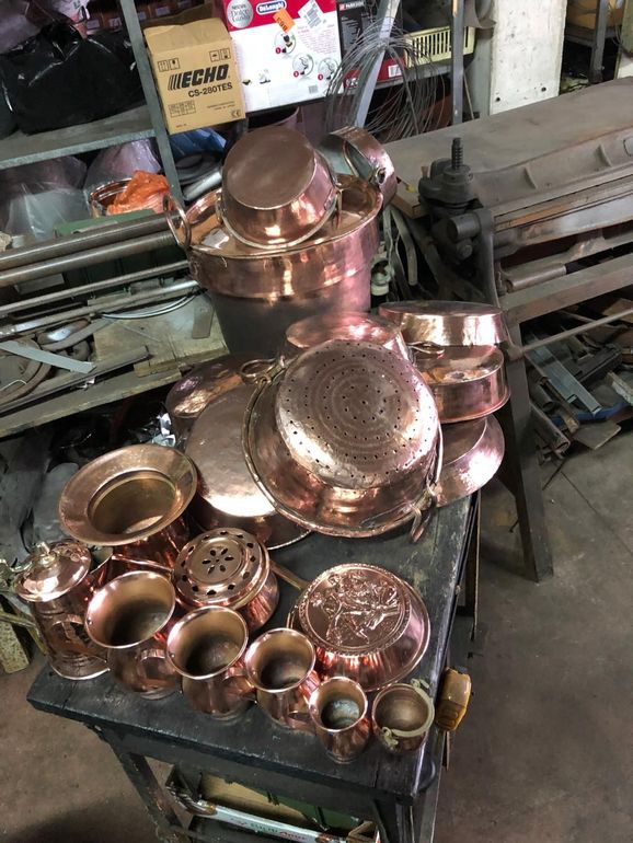 Una collezione di pentole, scolapasta e piccoli contenitori in rame lucidato, appoggiati su un banco da lavoro in un'officina.