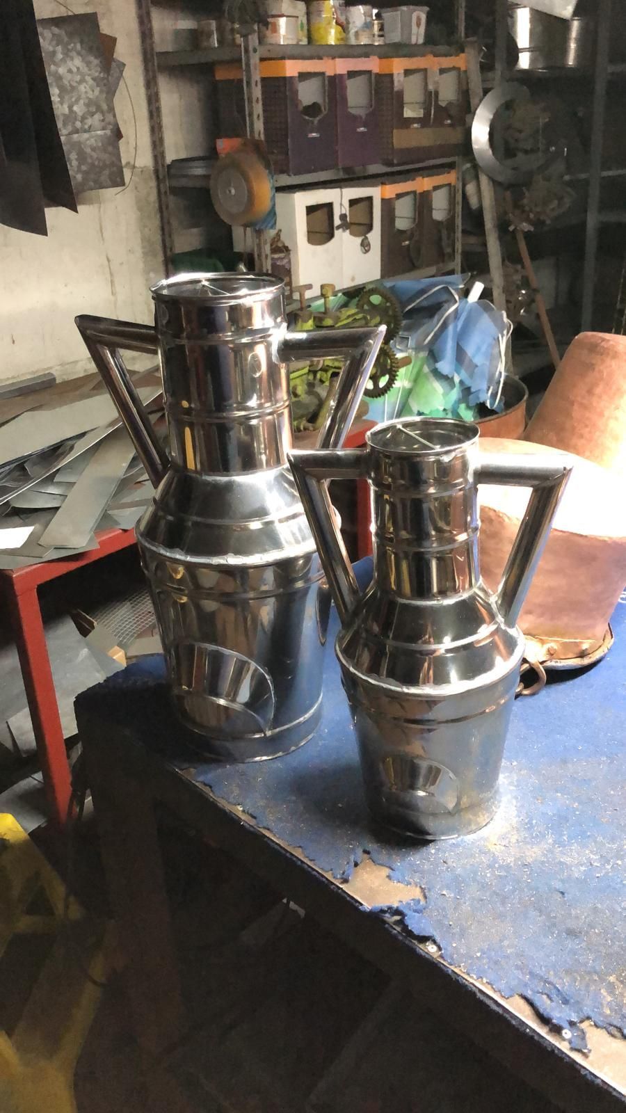 In un'officina disordinata, due contenitori di metallo lucido e brillante, dotati di manici, sono appoggiati su un banco da lavoro blu.