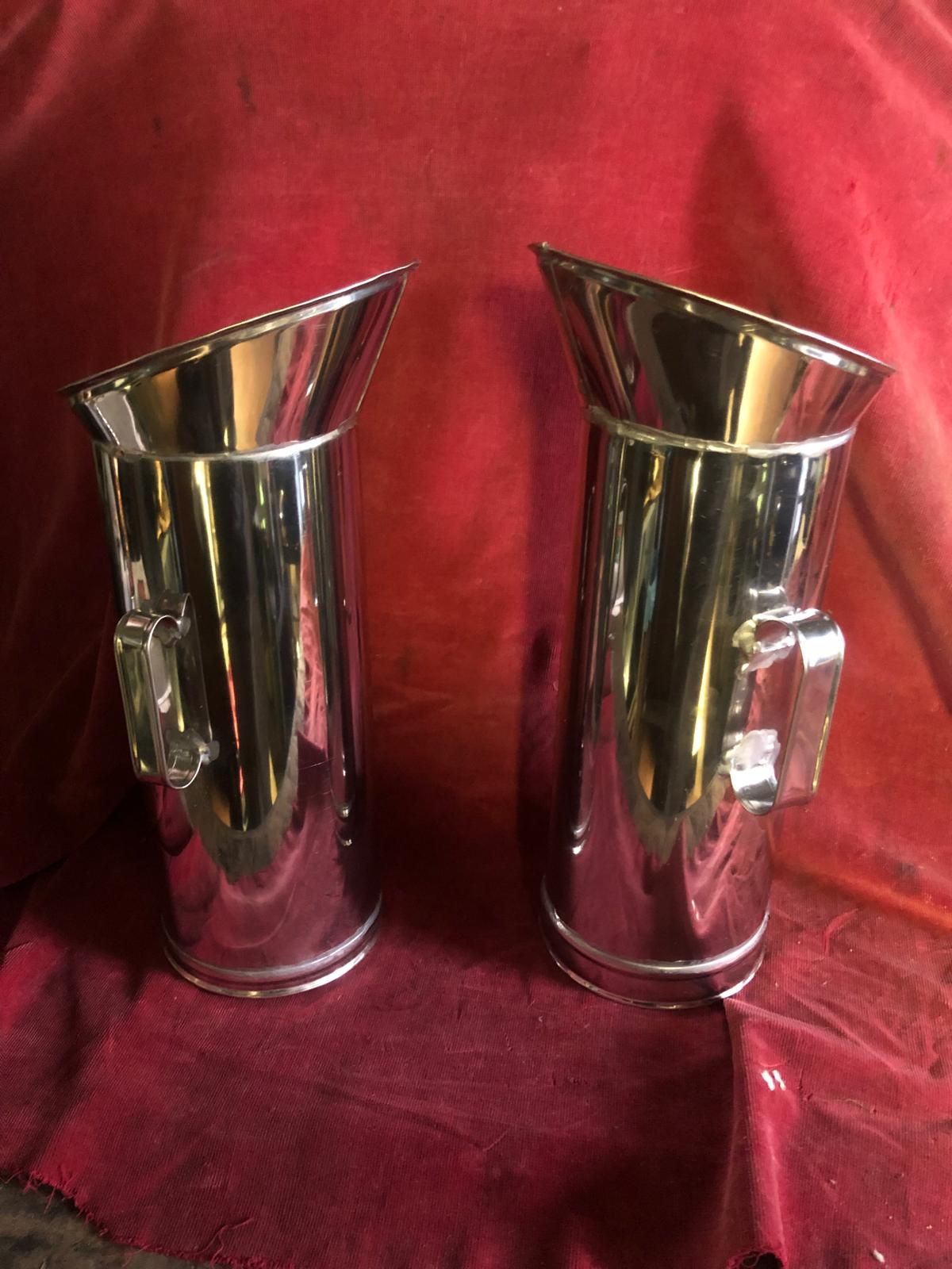Due brocche alte e cilindriche in metallo lucido, con beccucci angolati e manici laterali, poggiano su una superficie di tessuto rosso.