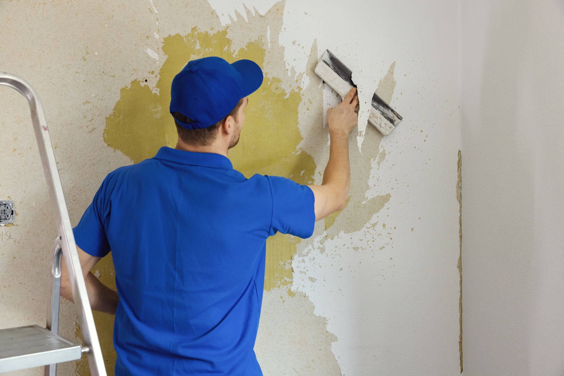 A worker diligently removing old painted wallpaper from the wall as part of a house renovation.