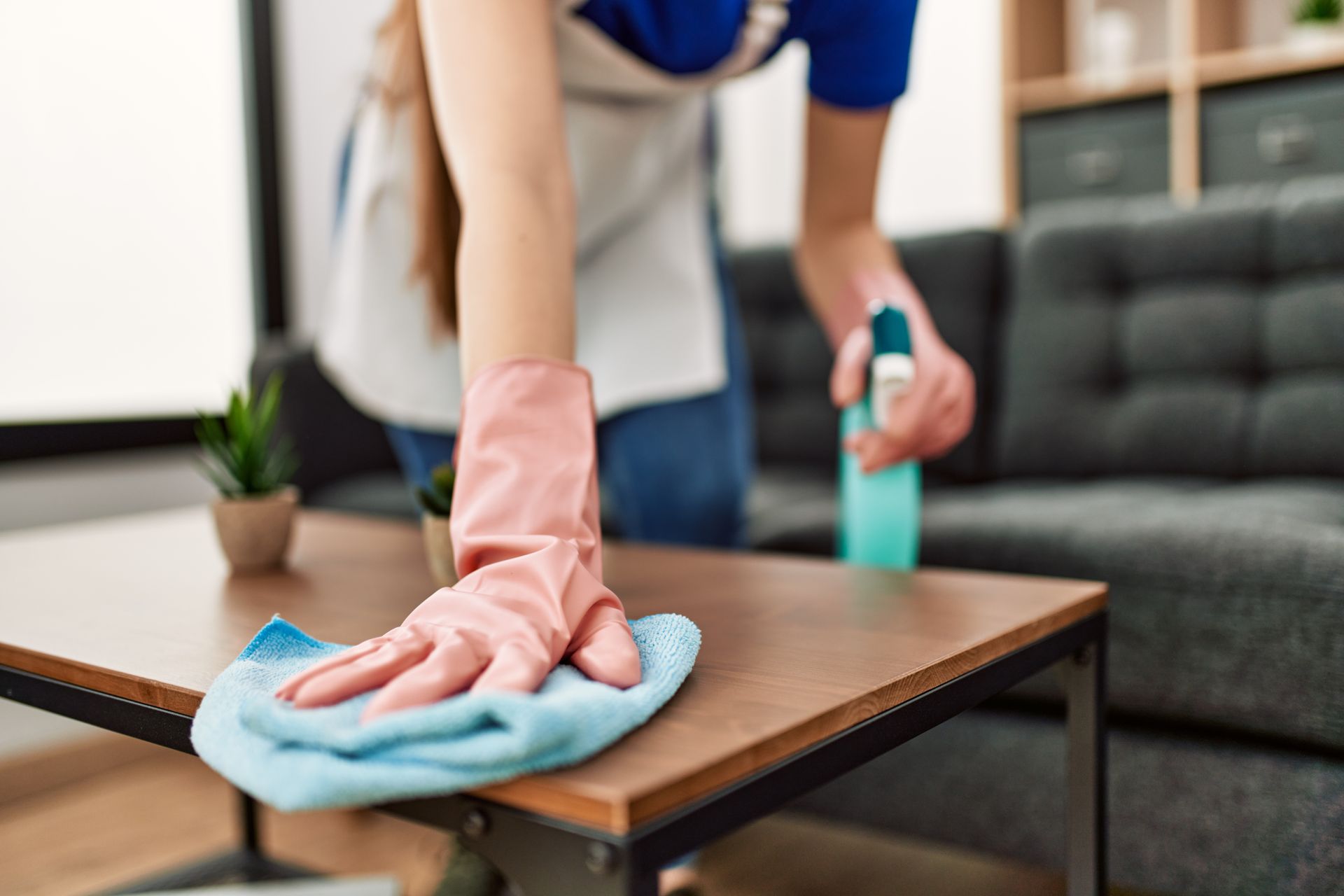 Person wiping a wooden table with a blue cloth and spray cleaner in a living room