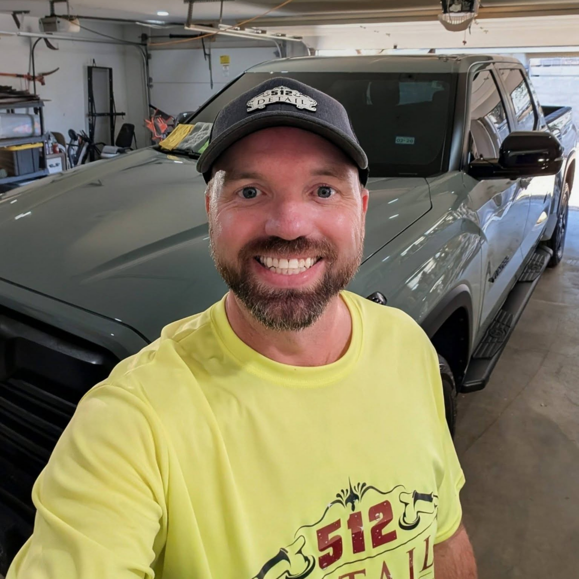 Smiling man in a neon yellow shirt and black cap taking a selfie in a garage beside a gray truck