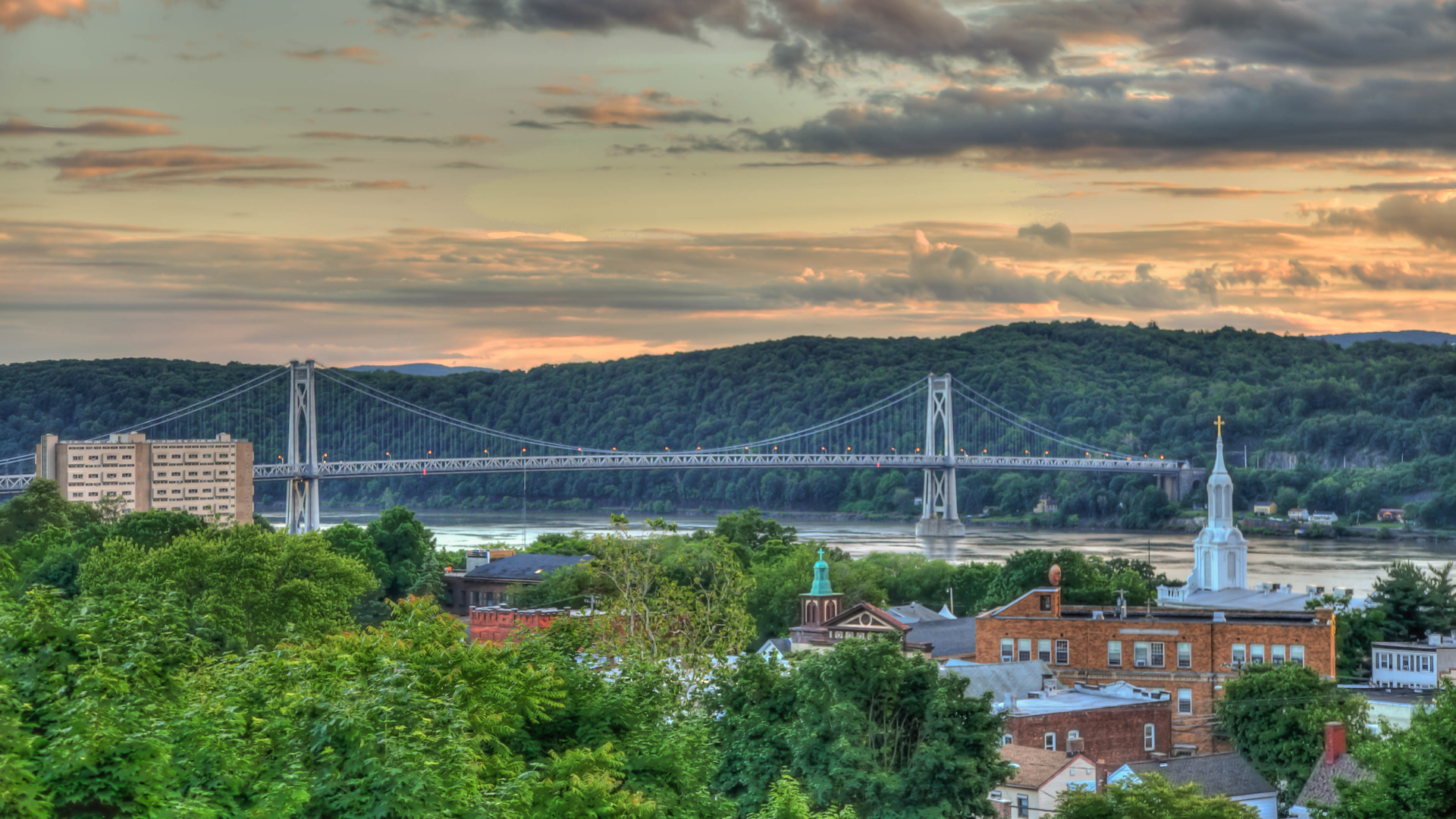 A scenic view of the Mid-Hudson Bridge spanning the Hudson River at sunset, overlooking a town with dense green trees.