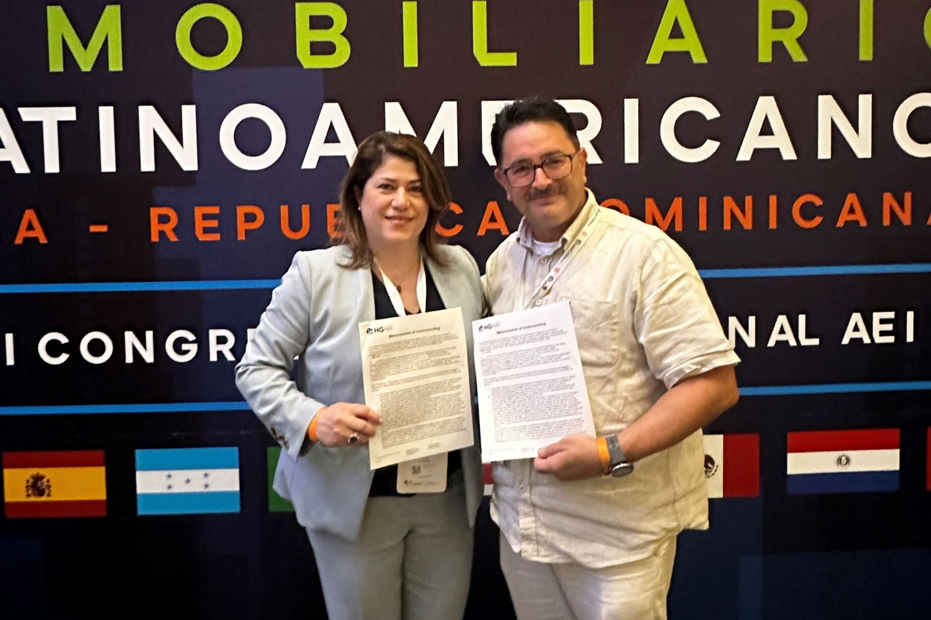 HGAR President Lynda Fernandez and CILA President Enrique Morales are holding papers, smiling, in front of a banner with text and flags.