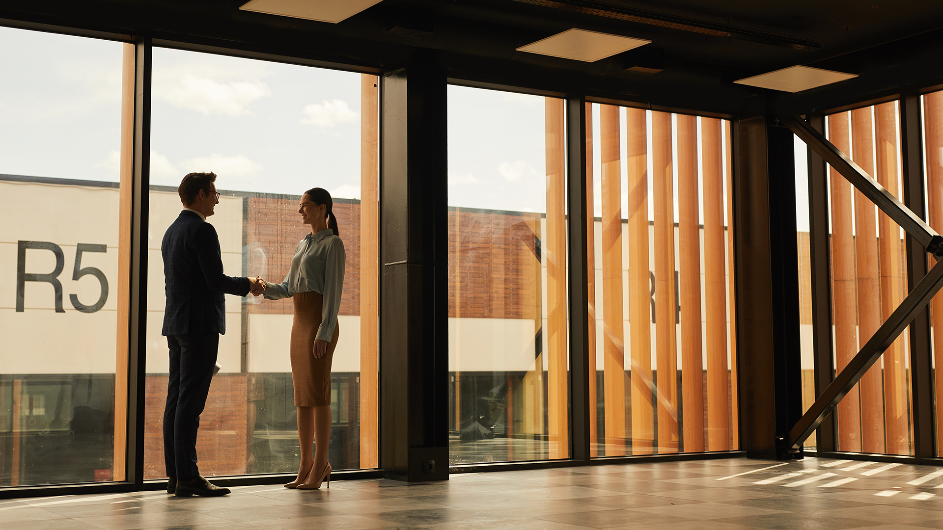 Man and woman shaking hands in a modern room with large windows and vertical wooden slats.
