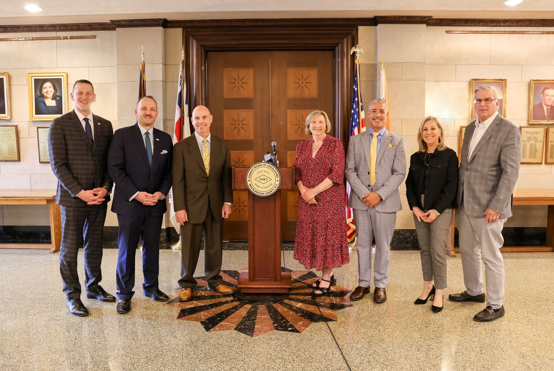 Seven people posing in a formal lobby around a pedestal clock.