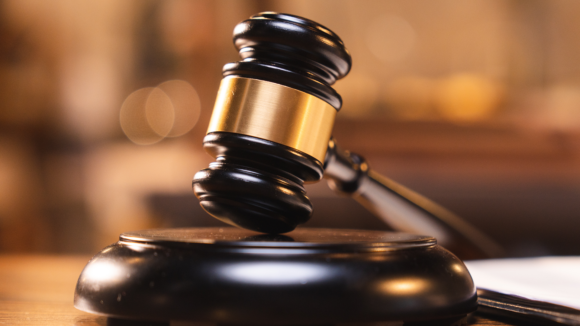 A wooden gavel rests on a round block in a softly lit, out-of-focus courtroom setting.